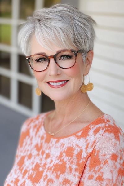 A woman with short, layered, silver-blonde hair styled with side-swept bangs, wearing glasses and gold earrings.