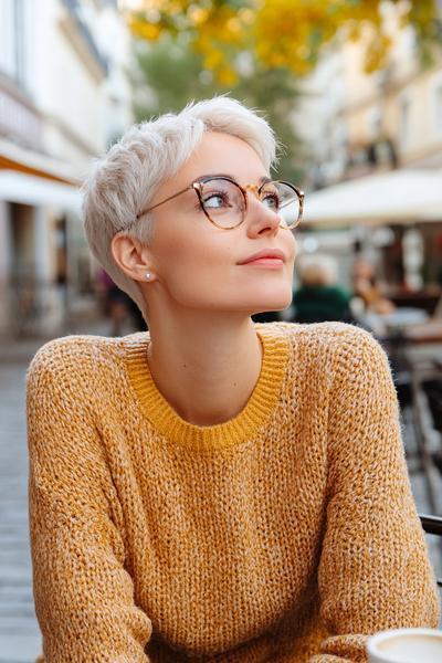 An older lady with glasses is wearing a sleek, platinum blonde pixie cut, complementing her stylish orange knit sweater.