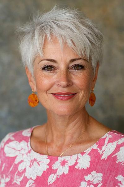 A woman with short, white, tousled pixie cut wearing floral pink top and orange earrings, showcasing a chic and easy-to-maintain wash and wear hairstyle for women over 60.