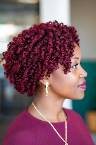 A profile view of a woman with short, tightly curled burgundy dark red hair, complementing her matching burgundy top and gold earrings.