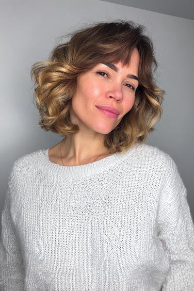 A woman with a chin-length, wavy bob hairstyle featuring soft, wispy fringe and subtle blonde highlights.