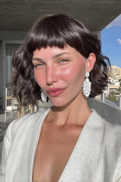 A woman with a wavy bob haircut featuring short, blunt fringe, styled in a glossy dark brown shade.