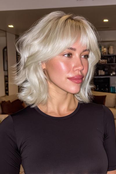A woman with platinum blonde, chin-length bob hair styled in loose waves, featuring a soft, wispy fringe.
