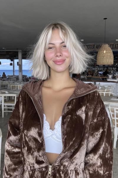 A young woman with platinum blonde, chin-length bob hair featuring soft, wispy fringe stands in a casual indoor setting.