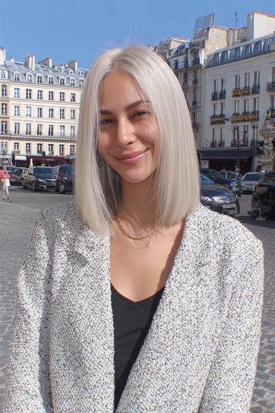 A woman with sleek, straight platinum blonde hair styled in a chin-length blunt bob, parted in the middle, standing outdoors in a city setting.