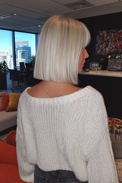 A woman with straight, platinum blonde hair styled in a blunt bob with a full fringe, seen from behind in an office setting.