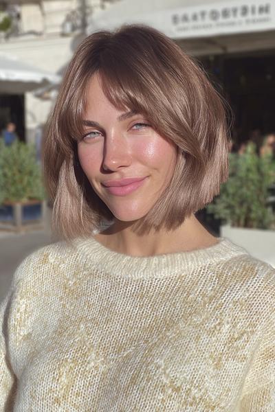 A woman with a chin-length blunt bob hairstyle featuring soft, wispy fringe, with light brown hair and subtle highlights.