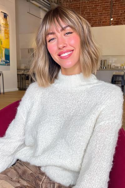 A woman with a chin-length bob haircut and soft, wispy fringe, featuring slightly wavy, highlighted hair.