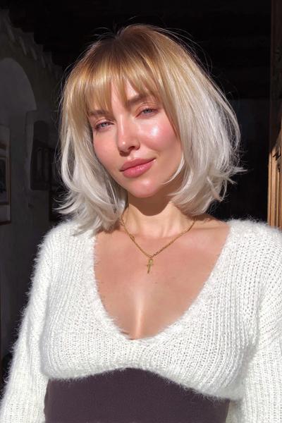 A woman with a chin-length bob haircut featuring soft, blunt bangs and platinum blonde hair with darker roots.