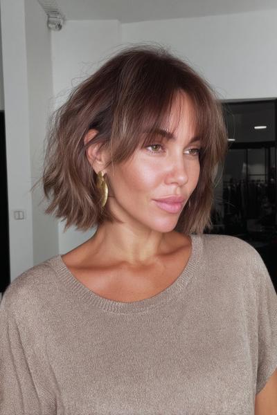A woman with a tousled, chin-length bob haircut and soft, wispy fringe, styled in a casual, textured look.