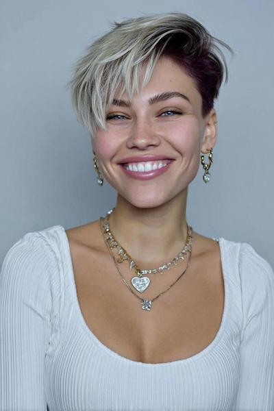 A woman with an undercut pixie hairstyle featuring dark roots and short sides, with longer, textured platinum blonde hair swept to one side.
