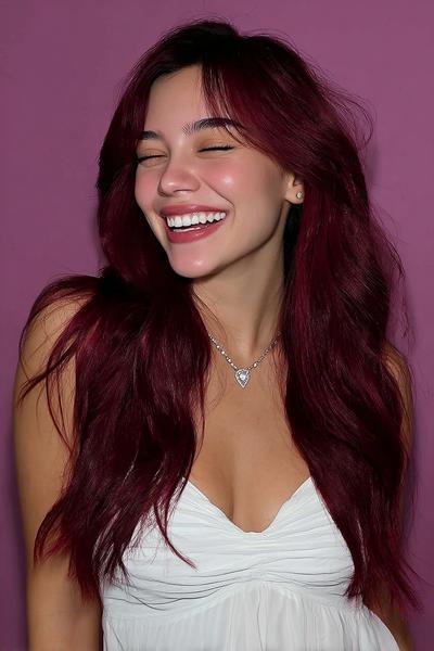 A young woman with long, layered, deep burgundy red hair styled in loose waves, wearing a white top and smiling against a purple background.