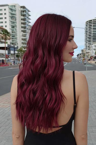 A woman with long, wavy, deep burgundy red hair is standing outdoors in an urban setting, wearing a black strappy top.