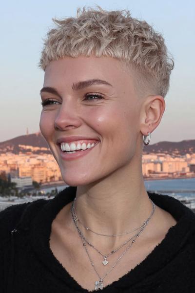 A woman with a short, textured blonde undercut pixie hairstyle featuring closely cropped sides and a choppy fringe, smiling outdoors.