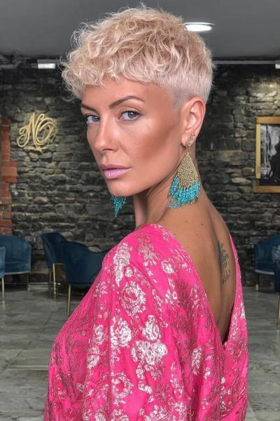 A woman with short, platinum blonde hair styled in a textured undercut pixie cut, with closely clipped sides and longer, softly curled hair on top.