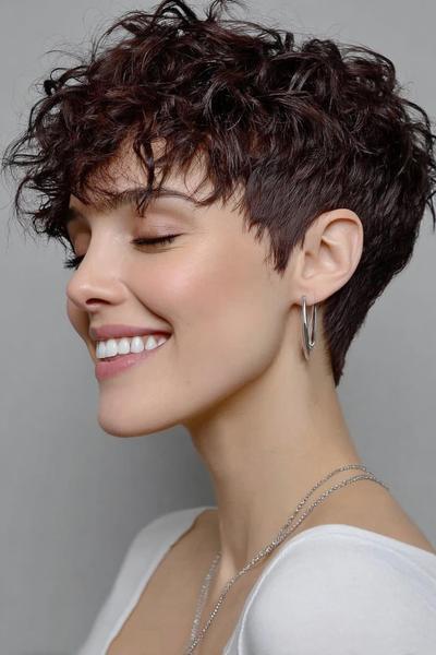 A woman with a short undercut pixie haircut featuring textured, curly layers on top and closely cropped sides, shown in profile.
