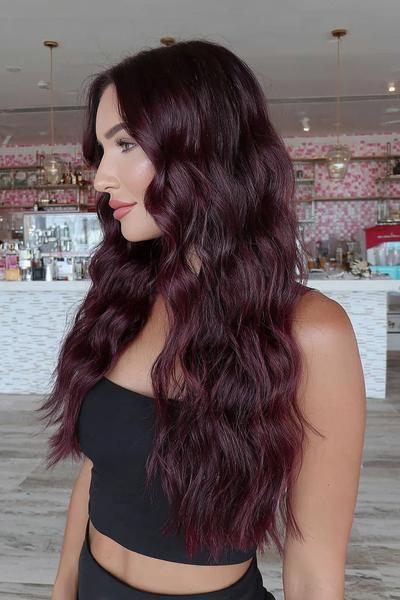 A woman with long, wavy, deep burgundy red hair is styled with a centre parting, wearing a black sleeveless top in a modern, well-lit interior.