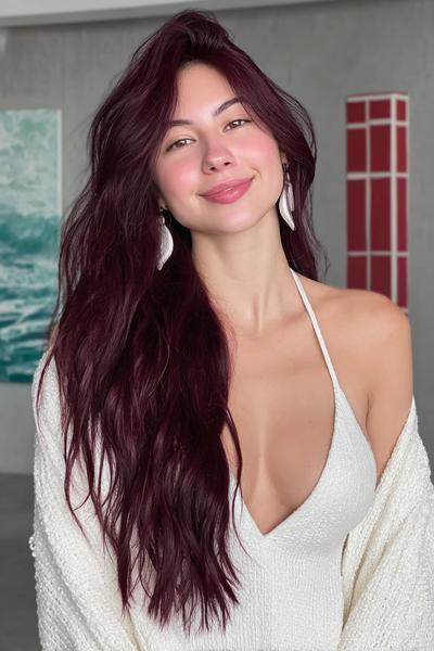 A woman with long, wavy, deep burgundy red hair is wearing a white sleeveless top and a cream cardigan.