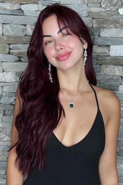 A woman with long, wavy deep burgundy red hair is wearing a black sleeveless top, silver earrings, and a necklace, standing in front of a stone wall.