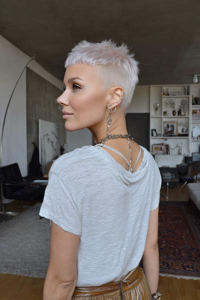 A woman with a platinum blonde undercut pixie hairstyle, featuring closely cropped sides and longer textured layers on top, is standing in a modern living room.