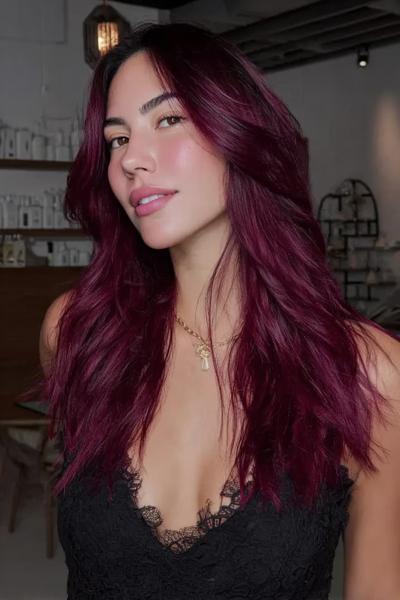 A woman with long, wavy, dark red hair styled with a middle parting is posing indoors.