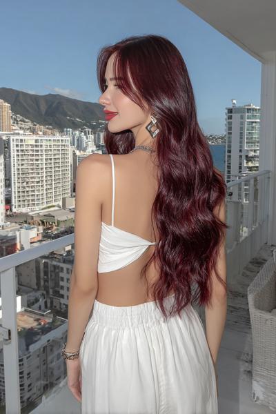 A woman with long, wavy, deep red hair is standing on a balcony, wearing a white backless top and skirt, with a cityscape and mountains in the background.