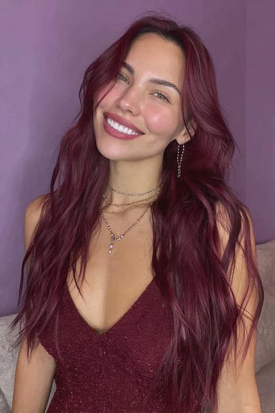 A woman with long, wavy, deep red hair styled in loose waves is smiling and wearing a matching burgundy dress.