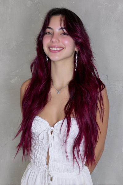 A young woman with long, wavy, vibrant burgundy red hair styled with curtain bangs is smiling in front of a plain grey background.