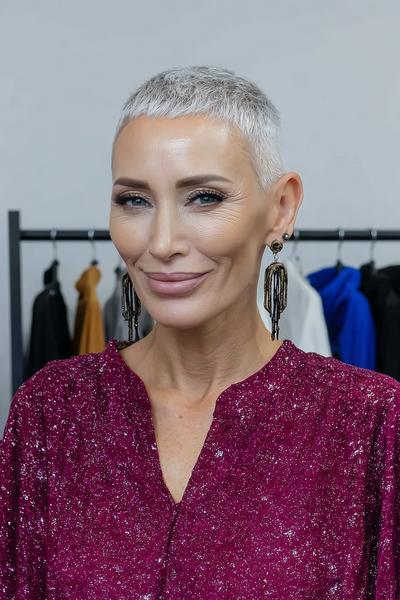 A woman with a short, textured, silver-grey undercut pixie hairstyle is wearing a sparkly burgundy top and statement earrings, standing indoors.