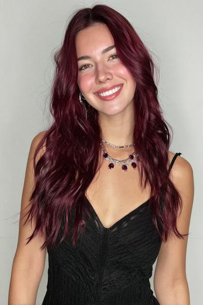 A woman with long, wavy, deep red hair styled in loose waves, wearing a black sleeveless dress and layered necklaces, smiles against a plain background.