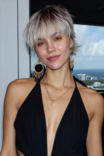 A young woman with a textured, platinum blonde undercut pixie hairstyle, featuring choppy layers and darker roots, is wearing statement earrings and a black halter top.