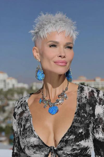 A woman with a short, textured, silver undercut pixie hairstyle is wearing statement blue earrings and a patterned top.