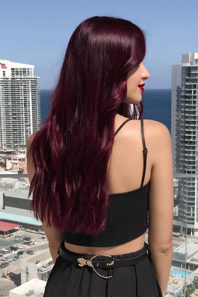 A woman with long, wavy, deep burgundy red hair is standing on a balcony overlooking a cityscape and the sea.