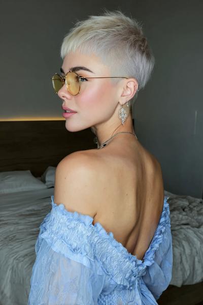 A woman with a platinum blonde undercut pixie hairstyle, featuring closely shaved sides and longer textured layers on top, is wearing round sunglasses and a light blue off-the-shoulder dress.