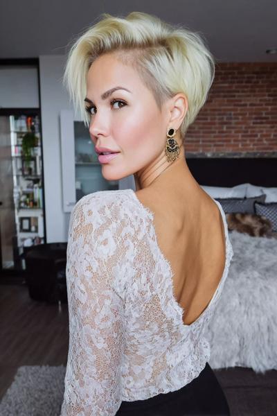 A woman with a platinum blonde undercut pixie hairstyle, featuring longer textured layers on top and closely shaved sides, is looking over her shoulder.