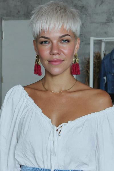 A woman with platinum blonde hair styled in a textured undercut pixie cut with choppy layers and a fringe, wearing statement red tassel earrings and a white off-the-shoulder top.