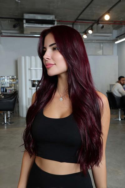 A woman with long, wavy, deep burgundy red hair styled with a centre parting is standing in a modern hair salon.