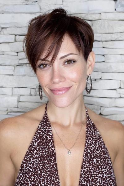 A woman with a stylish undercut pixie haircut featuring short sides and back with longer, side-swept layers on top, set against a white stone background.