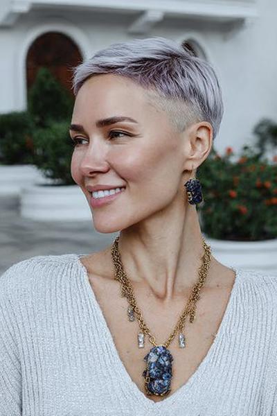 A woman with a stylish undercut pixie hairstyle, featuring closely cropped sides and longer, textured hair on top, is smiling outdoors.