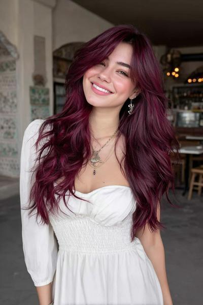 A woman with long, wavy deep red hair featuring voluminous layers and a centre parting, wearing a white dress and smiling indoors.
