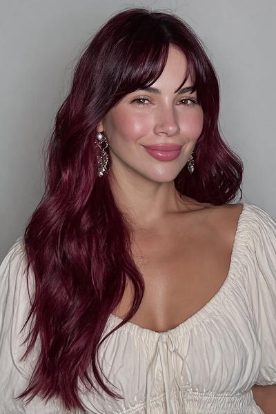 A woman with long, wavy deep red hair and soft curtain fringe is wearing a cream off-shoulder top and statement earrings.