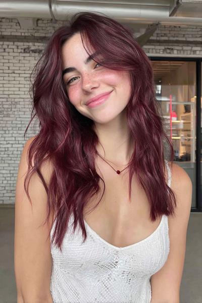 A young woman with long, wavy, deep red hair and soft curtain bangs is wearing a white sleeveless top and smiling indoors.