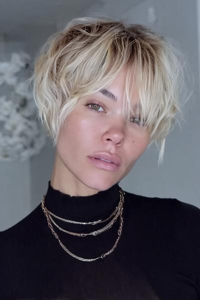 A woman with a tousled, blonde undercut pixie hairstyle featuring choppy layers and longer, textured fringe, wearing a black top and layered necklaces.