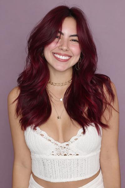 A woman with long, wavy, dark red hair styled with a centre parting is smiling at the camera.