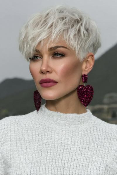A woman with platinum blonde hair styled in a textured, tousled undercut pixie hairstyle wears a white knitted jumper and statement heart-shaped earrings.