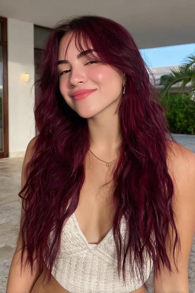 A young woman with long, wavy dark red hair styled with loose waves and soft, wispy fringe is wearing a white knitted top.