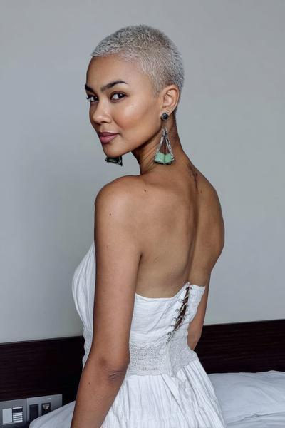 A woman with a closely-cropped, platinum blonde undercut pixie hairstyle, wearing a strapless white dress and statement earrings, looks over her shoulder.