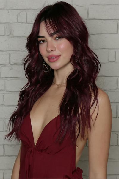 A young woman with long, wavy, dark red hair styled in loose waves with soft, wispy fringe, wearing a sleeveless burgundy dress.