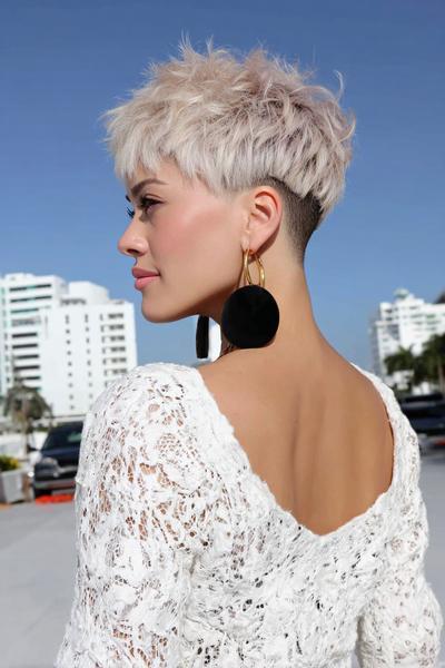A woman with platinum blonde hair styles an undercut pixie haircut, featuring longer, textured layers on top and closely shaved sides, photographed outdoors against a backdrop of modern buildings.