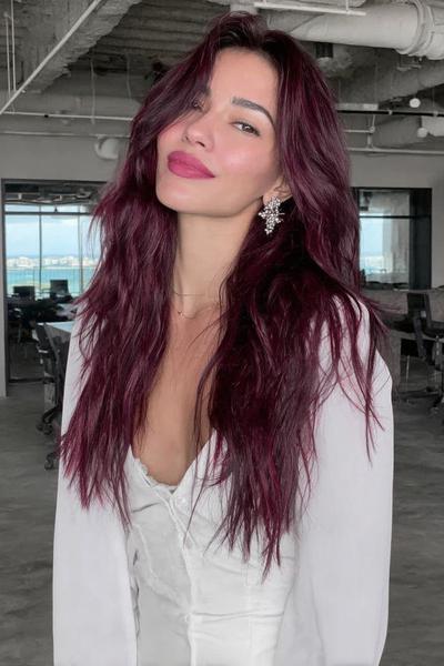 A woman with long, wavy, deep burgundy red hair is wearing a white blouse and standing indoors in an office setting.